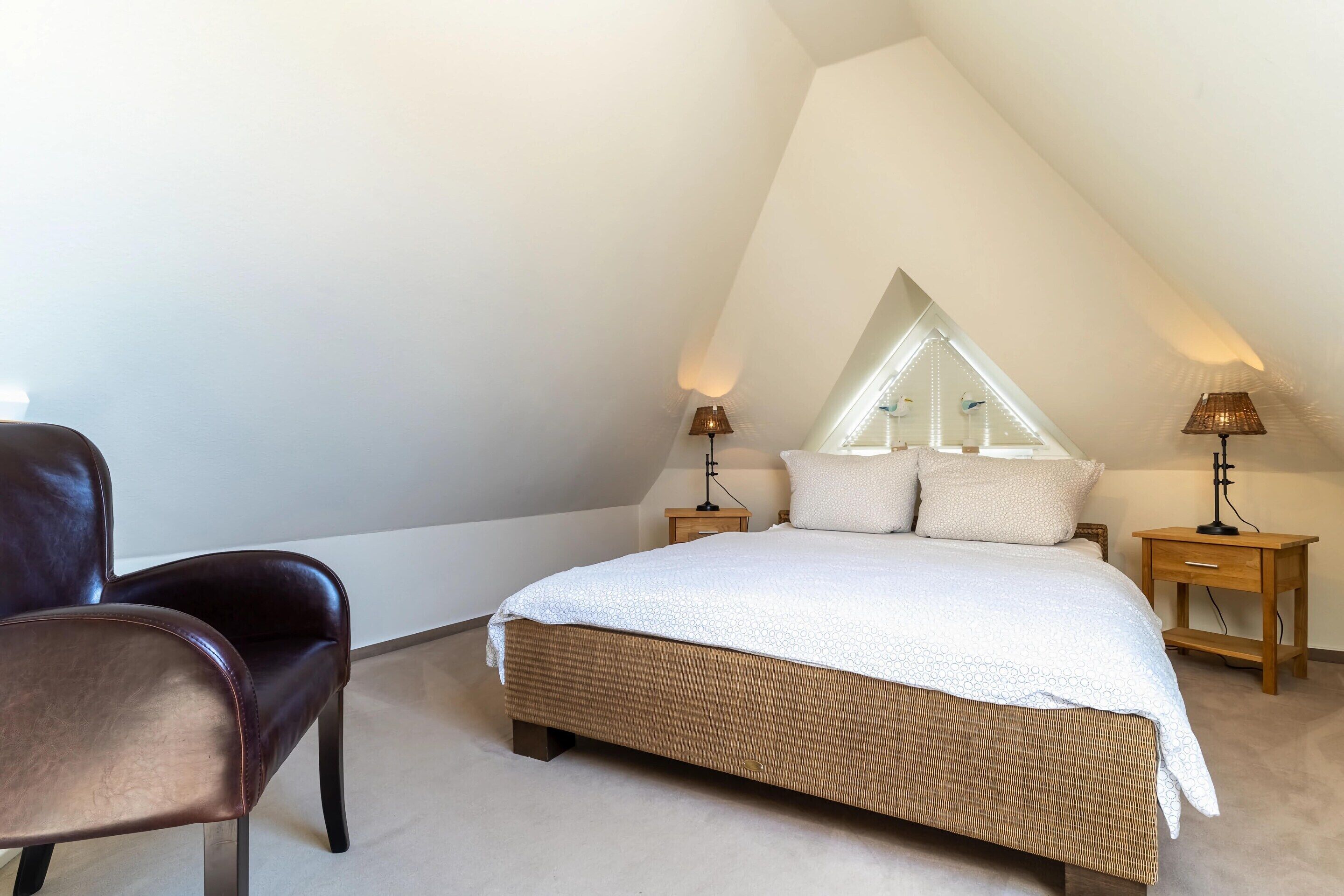 A bedroom with a sloped ceiling, a large bed with a white bedspread, two bedside tables with lamps, and a dark leather chair.