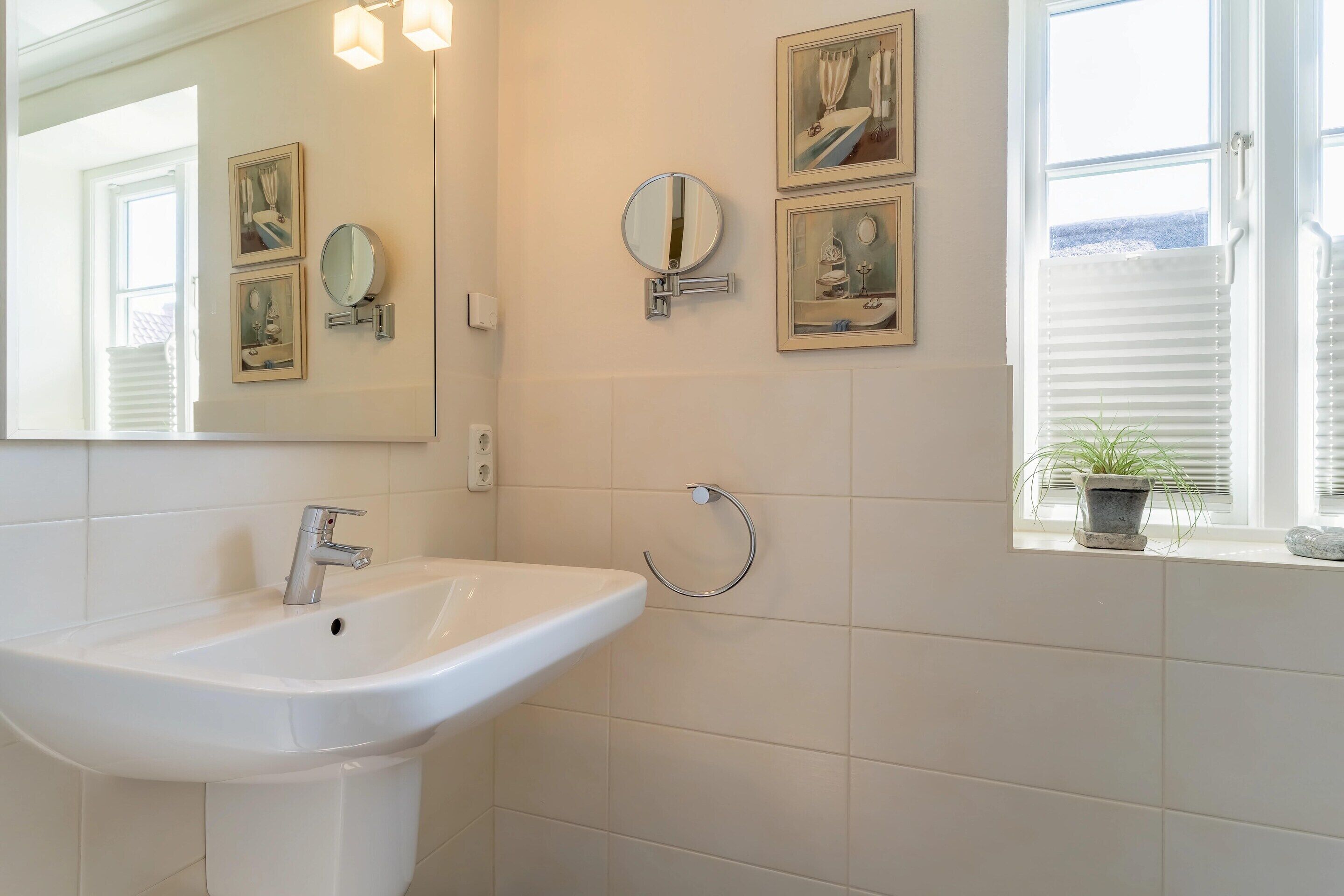 A bathroom with a white sink, two wall-mounted mirrors, framed artwork, and a window with blinds.