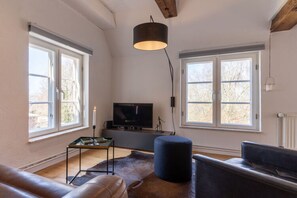 A modern living room with a leather sofa, a blue ottoman, a television, and a large window providing natural light. - Studio in Tonning Near sea (Toenning)
