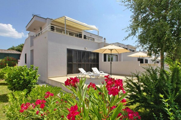 Une maison moderne à deux étages, dotée d’un patio couvert, de chaises longues blanches et d’un grand parasol.