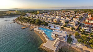 Luftaufnahme eines Küstenresorts mit großem Schwimmbecken, Villen direkt am Strand und einem Yachthafen.