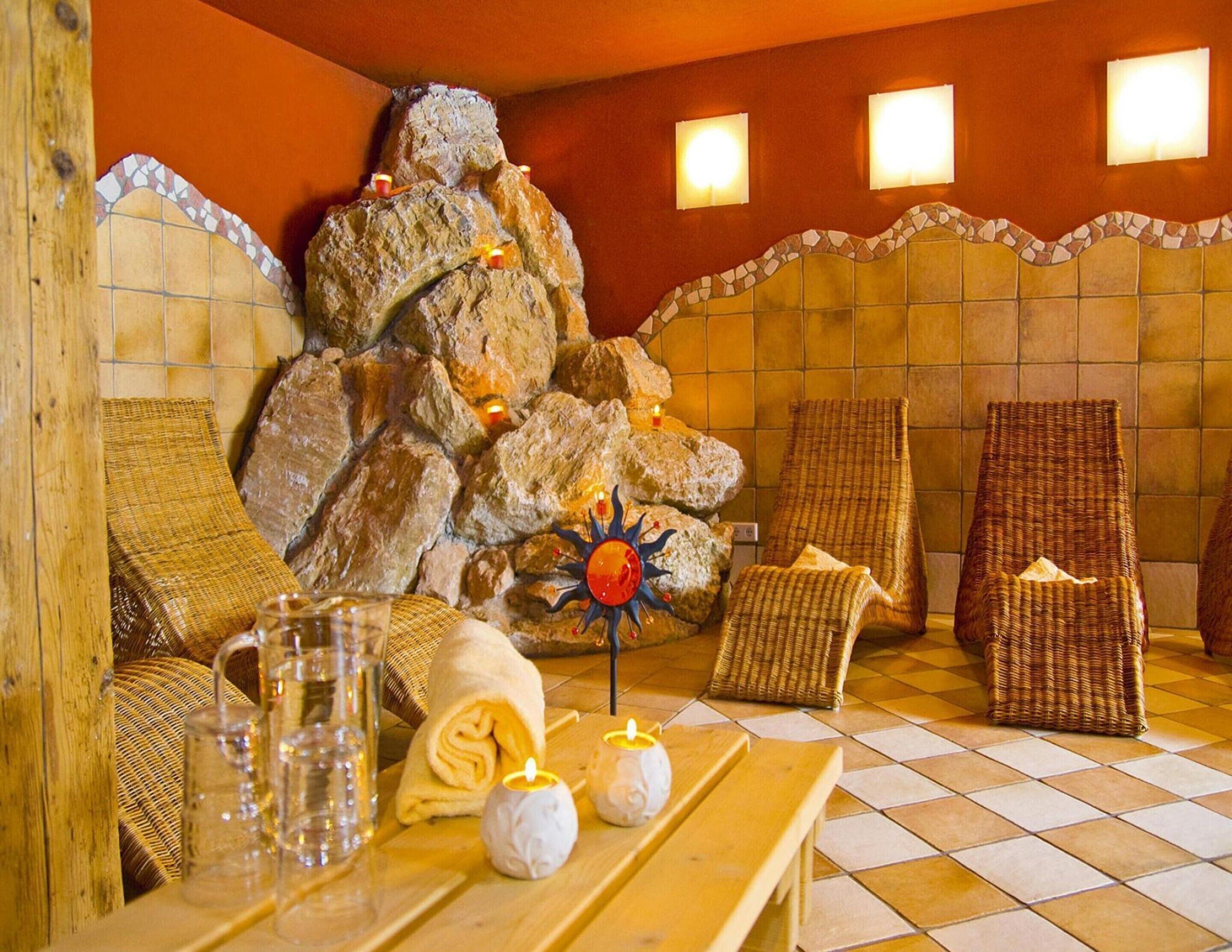A spa room with a stone wall, a sun decoration, wicker chairs, a wooden table with glasses and towels, and a tiled floor.