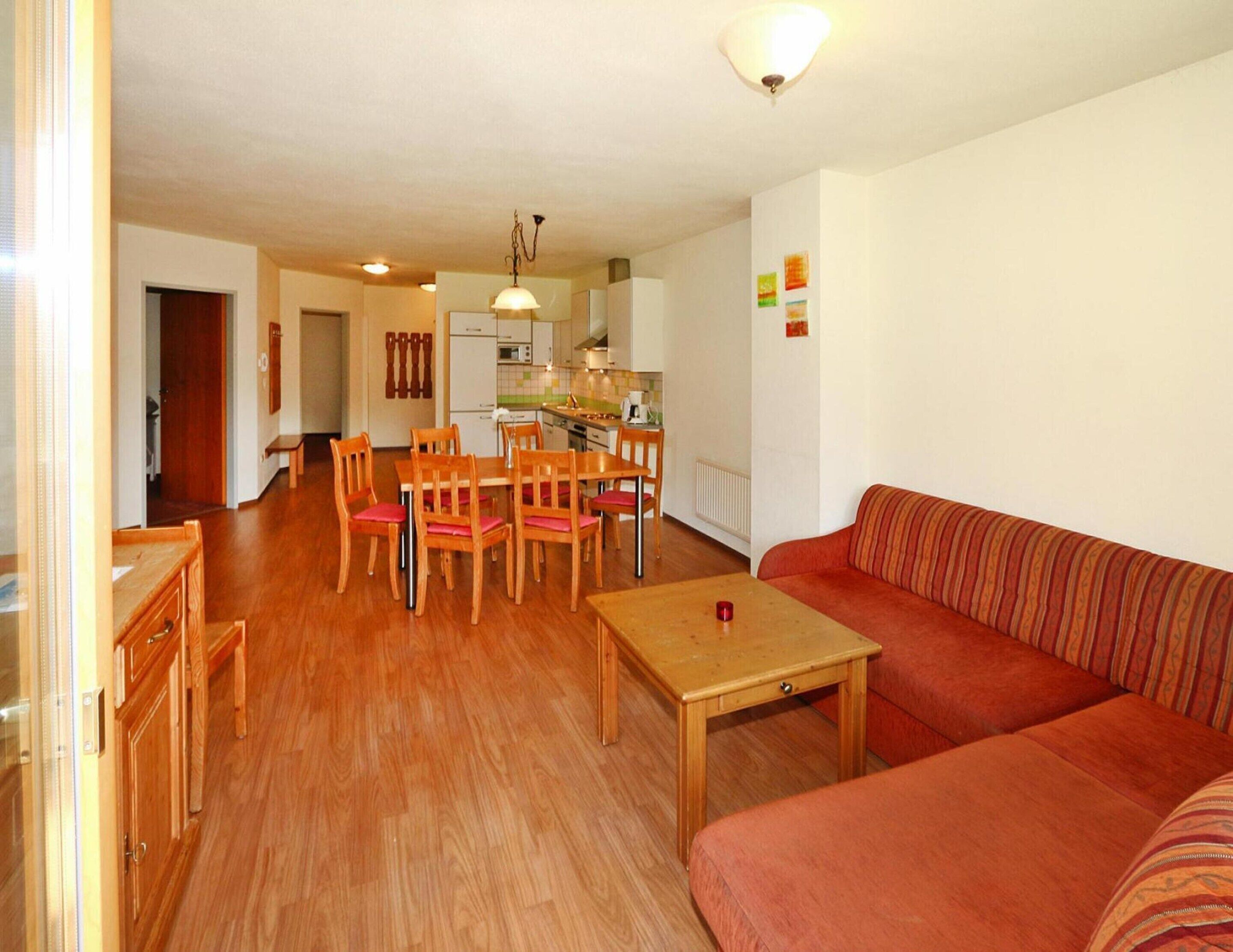A living room with a wooden floor, a red sofa, a dining table with chairs, and a kitchen in the background.
