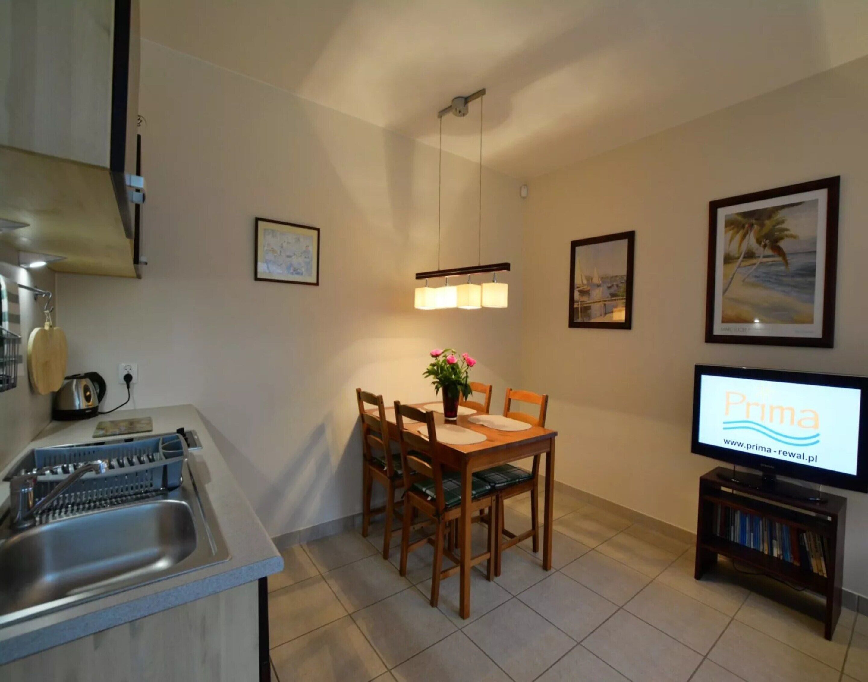 Een compacte keuken met een spoelbak, waterkoker en broodrooster. Er is een eethoek met een tafel en stoelen, een kleine televisie en beplakte foto’s aan de muur.