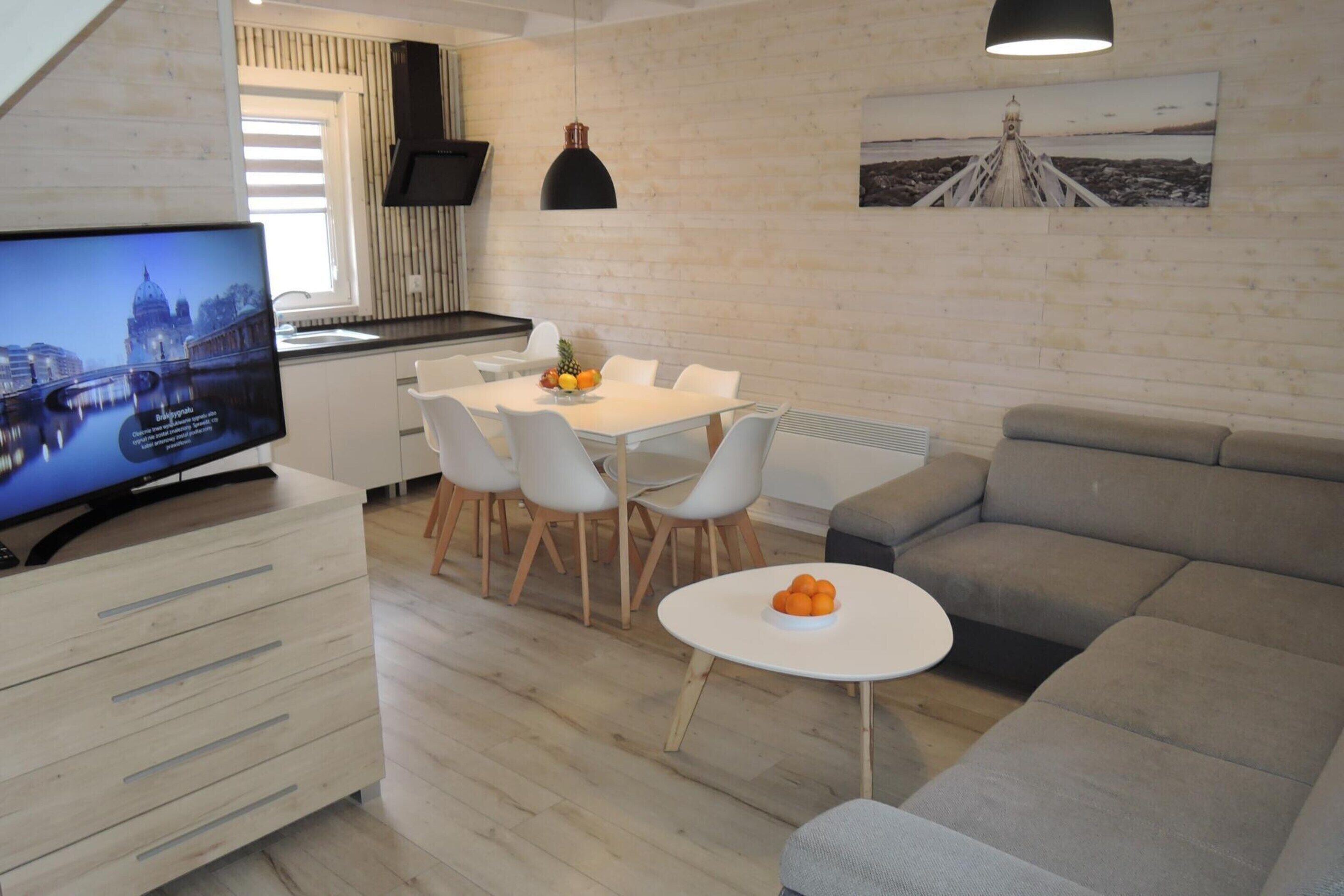 A modern living room with a TV, a dining area with chairs, a sofa, and a small table with fruit.