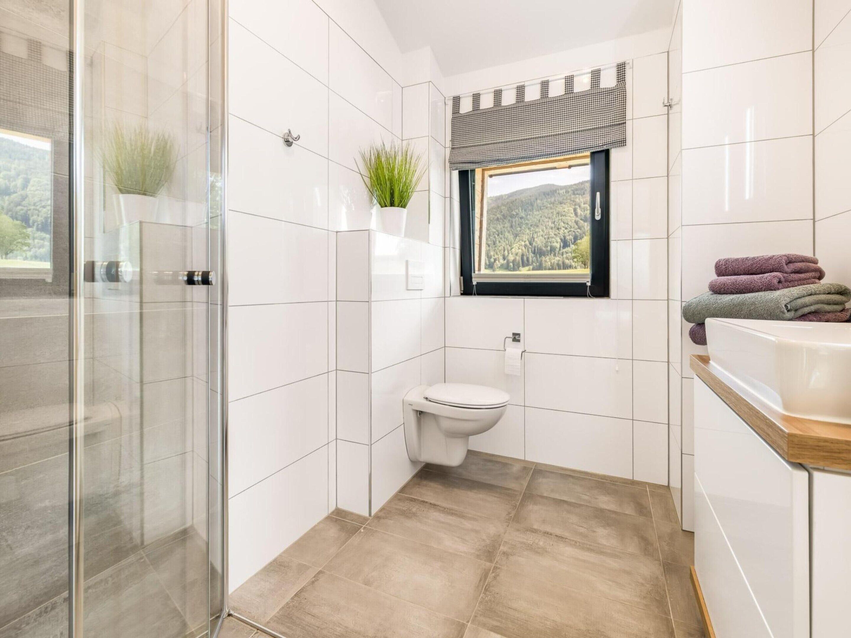 A bathroom with a glass shower enclosure, a toilet, a window with a view of a forest, and a sink with stacked towels.