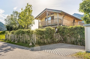 A wooden cabin with a balcony, surrounded by a wooden fence and greenery, with mountains in the background.