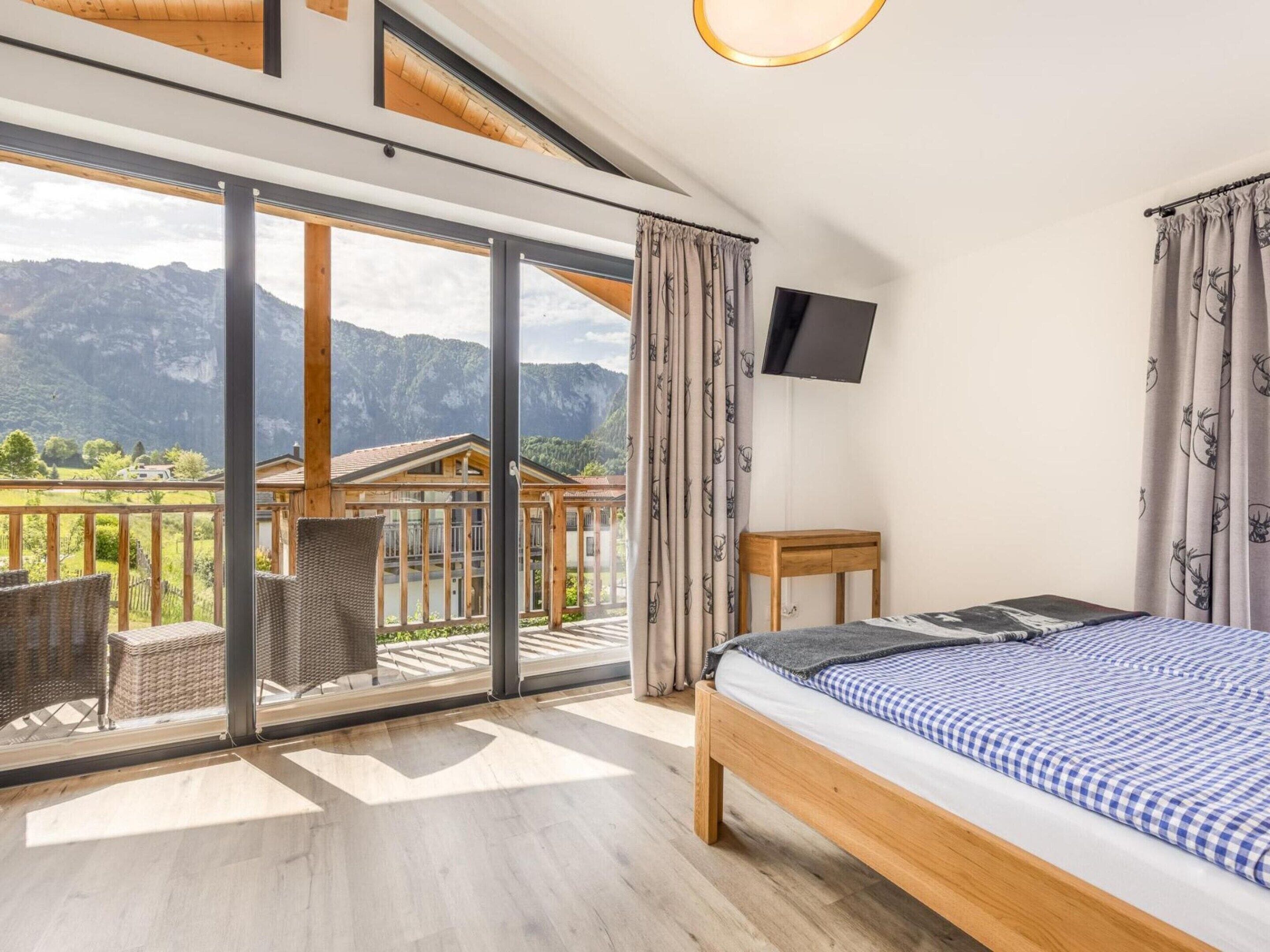 Un dormitorio con cama, televisor, balcón con mobiliario de mimbre y vistas a la montaña.
