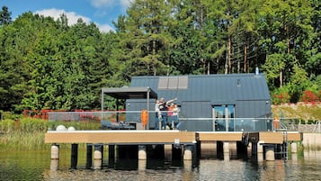 A modern houseboat with a wooden deck, a covered outdoor area, and a view of a forested lake.
