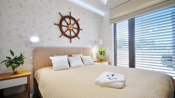 A hotel room with a large bed, a ship's wheel decoration, a potted plant, and a window with blinds.