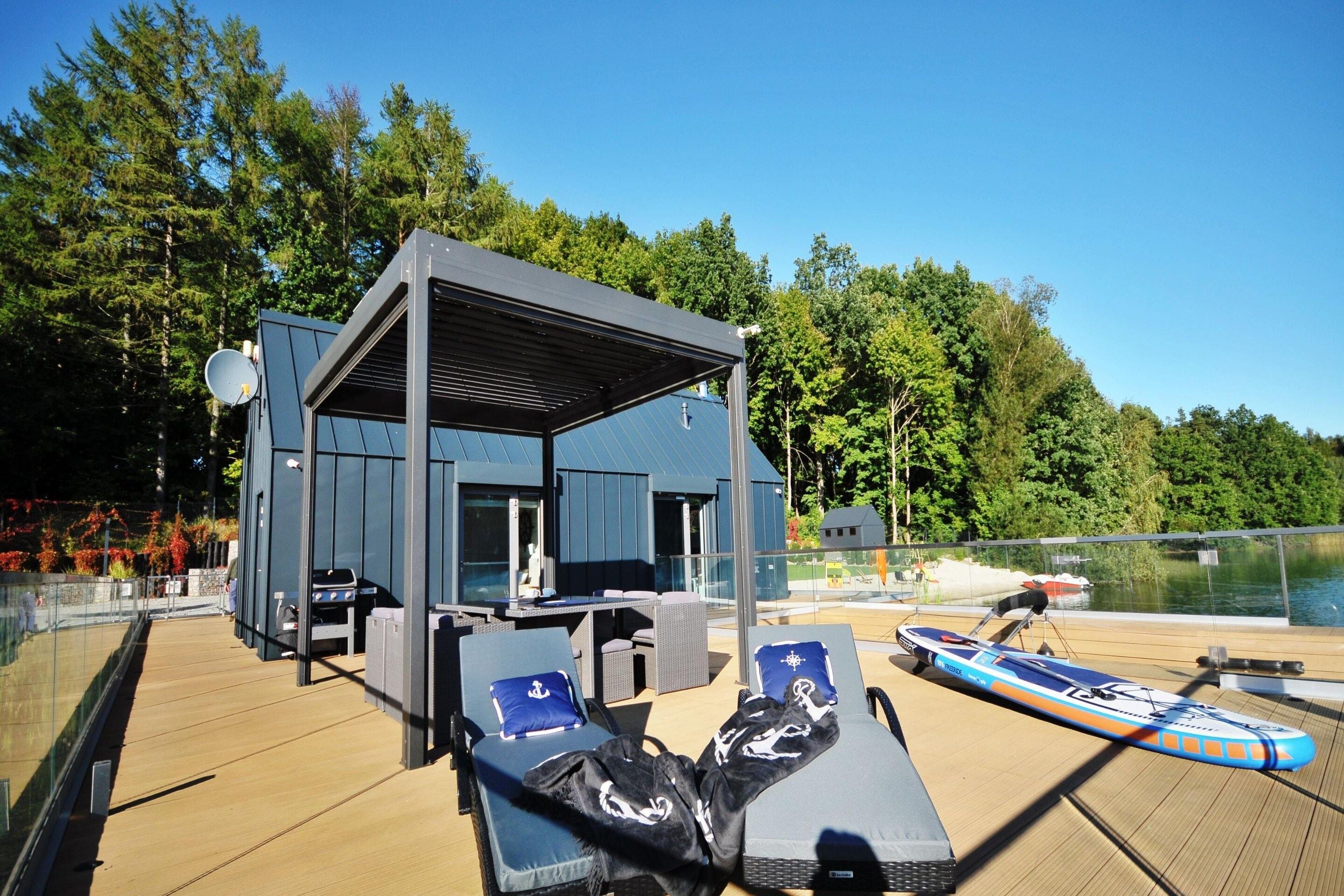 A covered outdoor seating area with lounge chairs and a barbecue grill, overlooking a body of water.