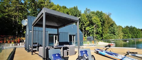 A covered outdoor seating area with lounge chairs and a barbecue grill, overlooking a body of water.