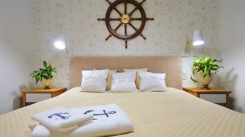 A hotel room with a bed, a ship's wheel decoration, two bedside tables with lamps, and two potted plants.