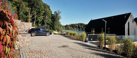 Une allée pavée mène à un bâtiment à l’architecture moderne, une voiture est garée devant, et on aperçoit une étendue d’eau en arrière-plan.