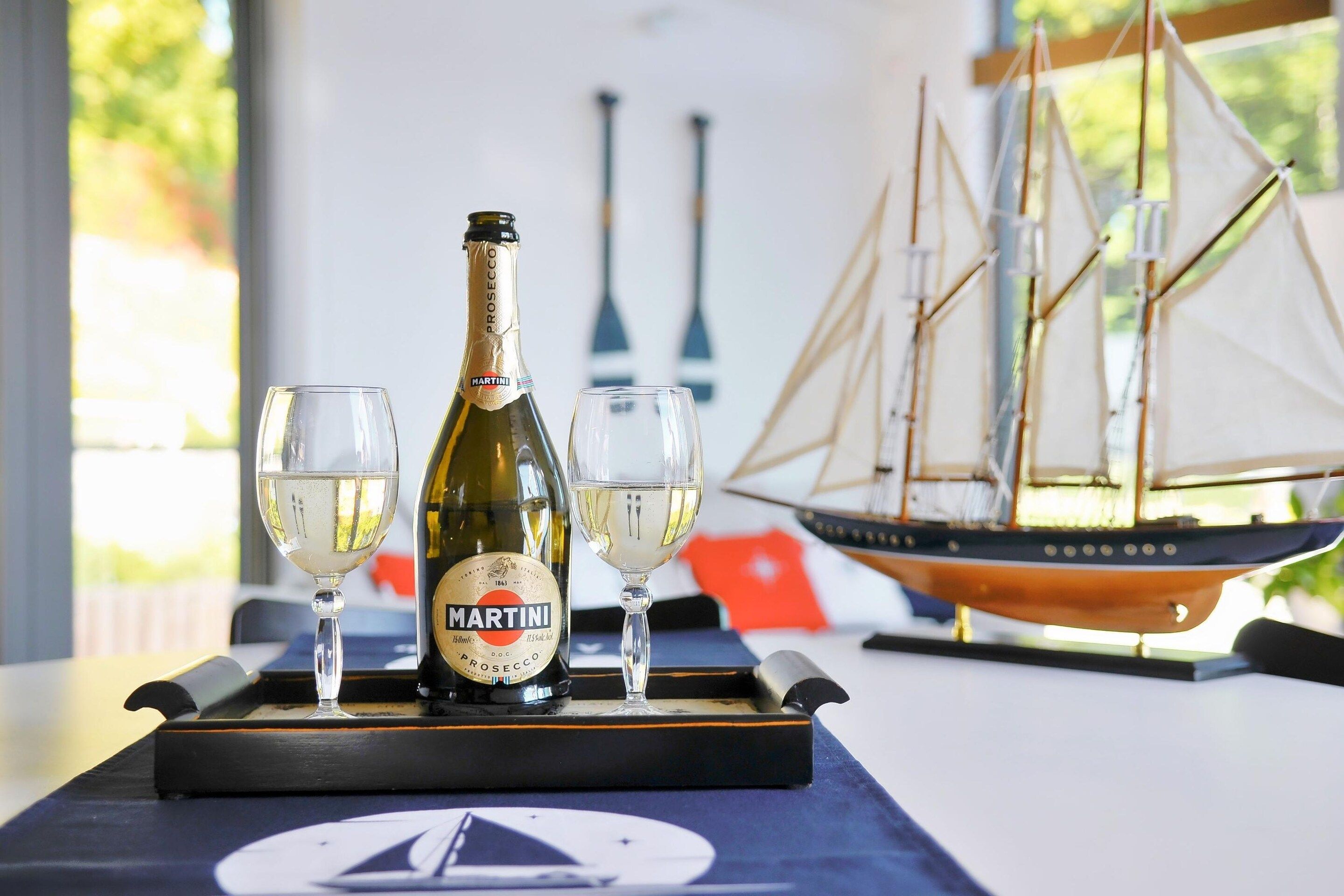 Una botella de Martini Prosecco sobre una bandeja con dos copas de vino, un barco modelo y un modelo de velero al fondo.