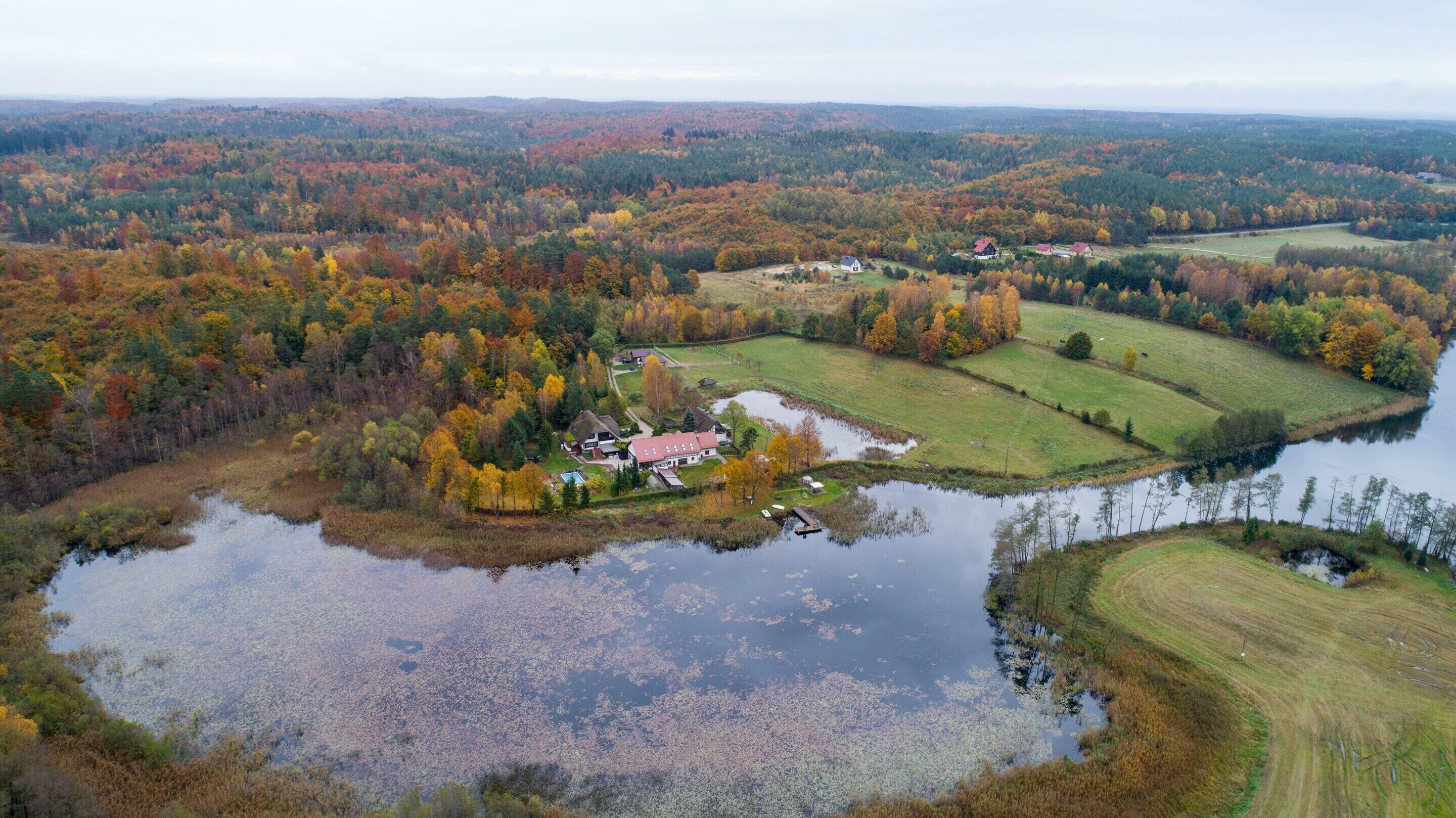 An aerial view of a rural area with a lake, surrounded by autumn-colored trees and a few houses.