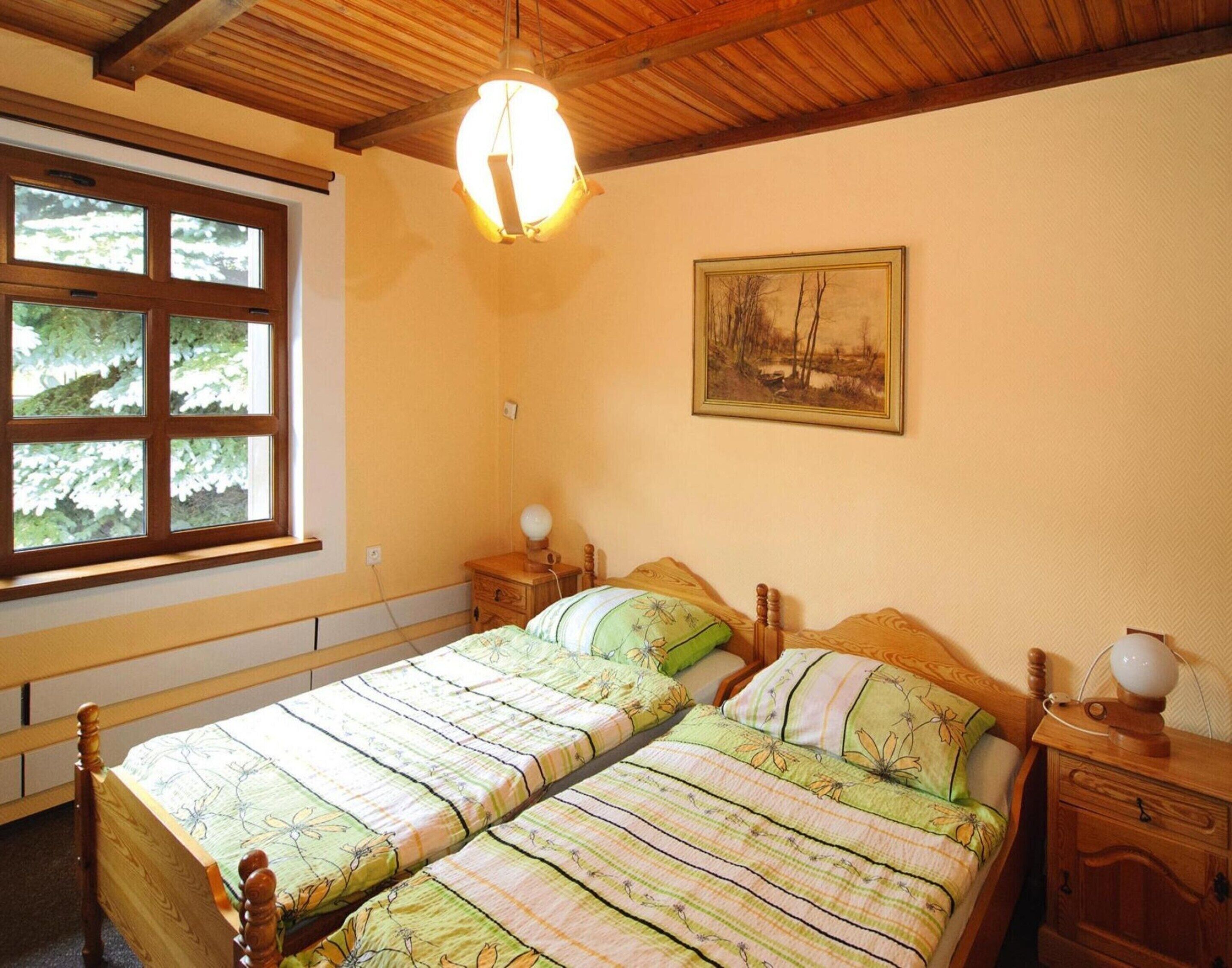 Une chambre avec deux lits simples, des têtes de lit en bois, une table de chevet avec une lampe, une fenêtre donnant sur des arbres et un tableau au mur.