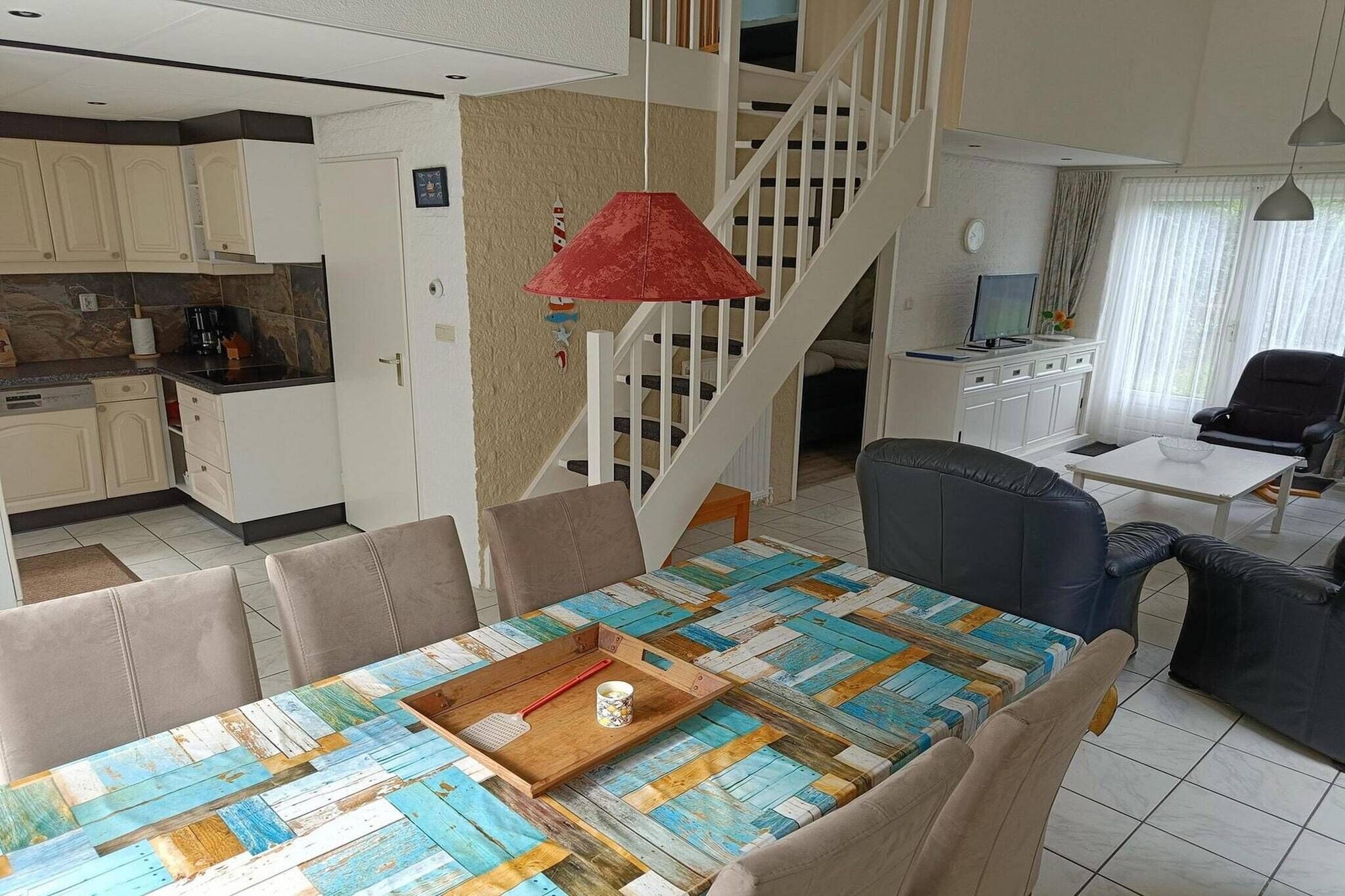 A dining area with a table set for four, a red hanging lamp, and a staircase leading to an upper level.
