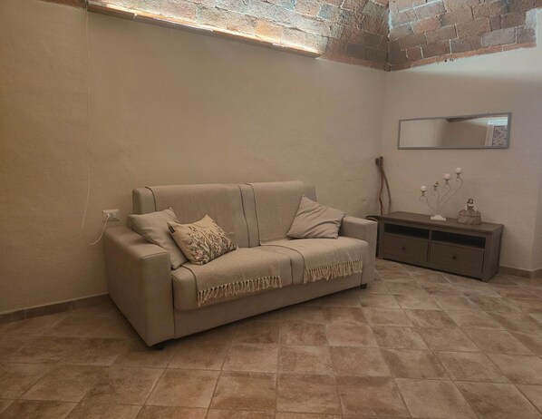 Un canapé beige agrémenté d’un plaid et de deux coussins, un meuble en bois orné d’objets décoratifs et un sol carrelé.