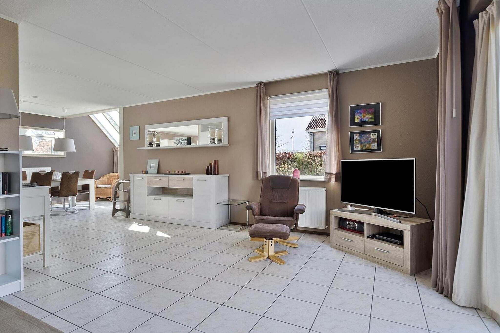 Un salon moderne doté d’un téléviseur à écran plat, d’un fauteuil confortable, d’un coin repas avec une table et des chaises, et d’une grande fenêtre agrémentée de rideaux.