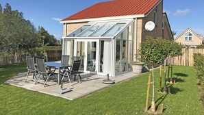 A house with a red roof, a glass conservatory, a patio with a table and chairs, and a garden.
