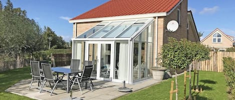 A house with a red roof, a glass conservatory, a patio with a table and chairs, and a garden.