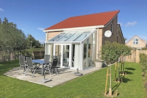 A house with a red roof, a glass conservatory, a patio with a table and chairs, and a garden.