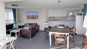 A modern living area with a sofa, coffee table, dining table, and chairs. There is a kitchen with white cabinets and a dining area with a table and chairs.