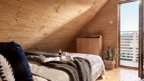 A cozy attic bedroom with wooden walls, a large bed with a knitted blanket, a bedside table with a book and a bottle, and a view of the outside through a sliding door. - Holiday Homes With Pool, Attractions for Children (Mielno)