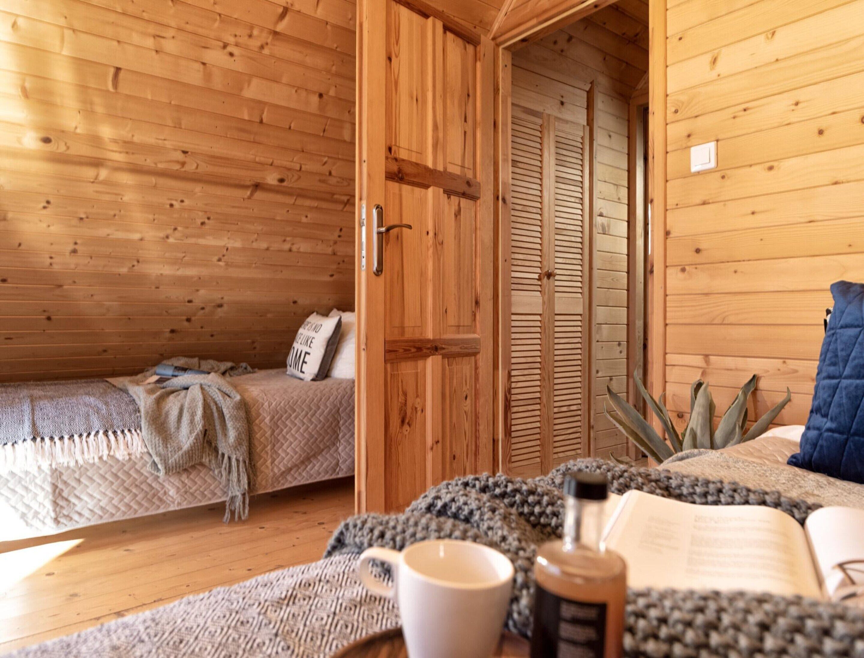 A cozy wooden room with a bed, a small table with a cup and a book, and a door leading to another room.