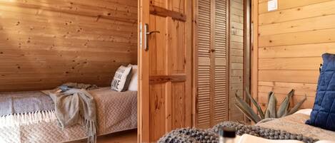 A cozy wooden room with a bed, a small table with a cup and a book, and a door leading to another room.