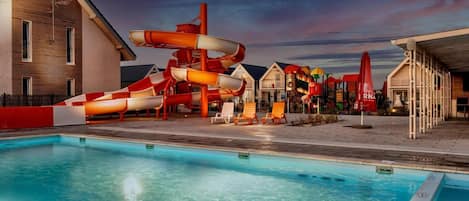 A pool area with a water slide, lounge chairs, and a building with a sloped roof.