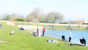 A lakeside setting with people fishing, a grassy area, and a building in the background.