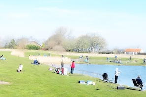 A lakeside setting with people fishing, a grassy area, and a building in the background.
