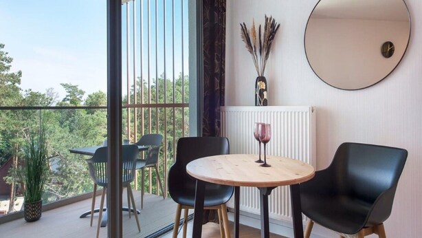 A modern room with a round table, two black chairs, a mirror, and a view of greenery through large windows. - Apartment in Pobierowo With SPA & Pools (Rewal)