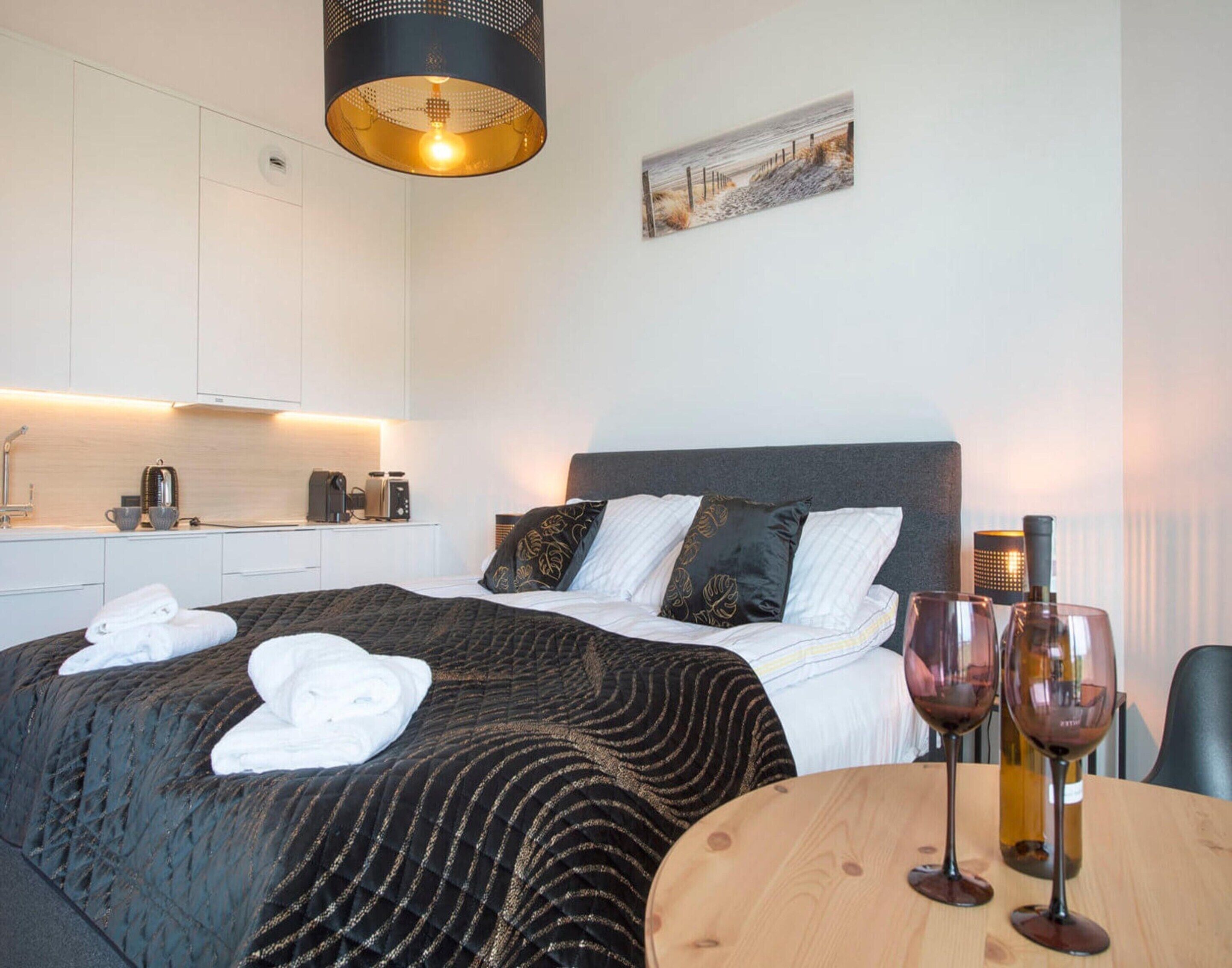 Moderni hotellihuone, jossa on sänky, pyöreä puinen pöytä viinilaseineen, seinällä taulu ja taustalla pieni keittiönurkkaus.