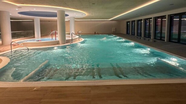 An indoor swimming pool with a central hot tub, surrounded by glass walls and a wooden deck.