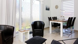 A modern dining area with a white table and black chairs, large windows with curtains, and a view of greenery outside. - Ferienhaus, Boiensdorf (Boiensdorf)