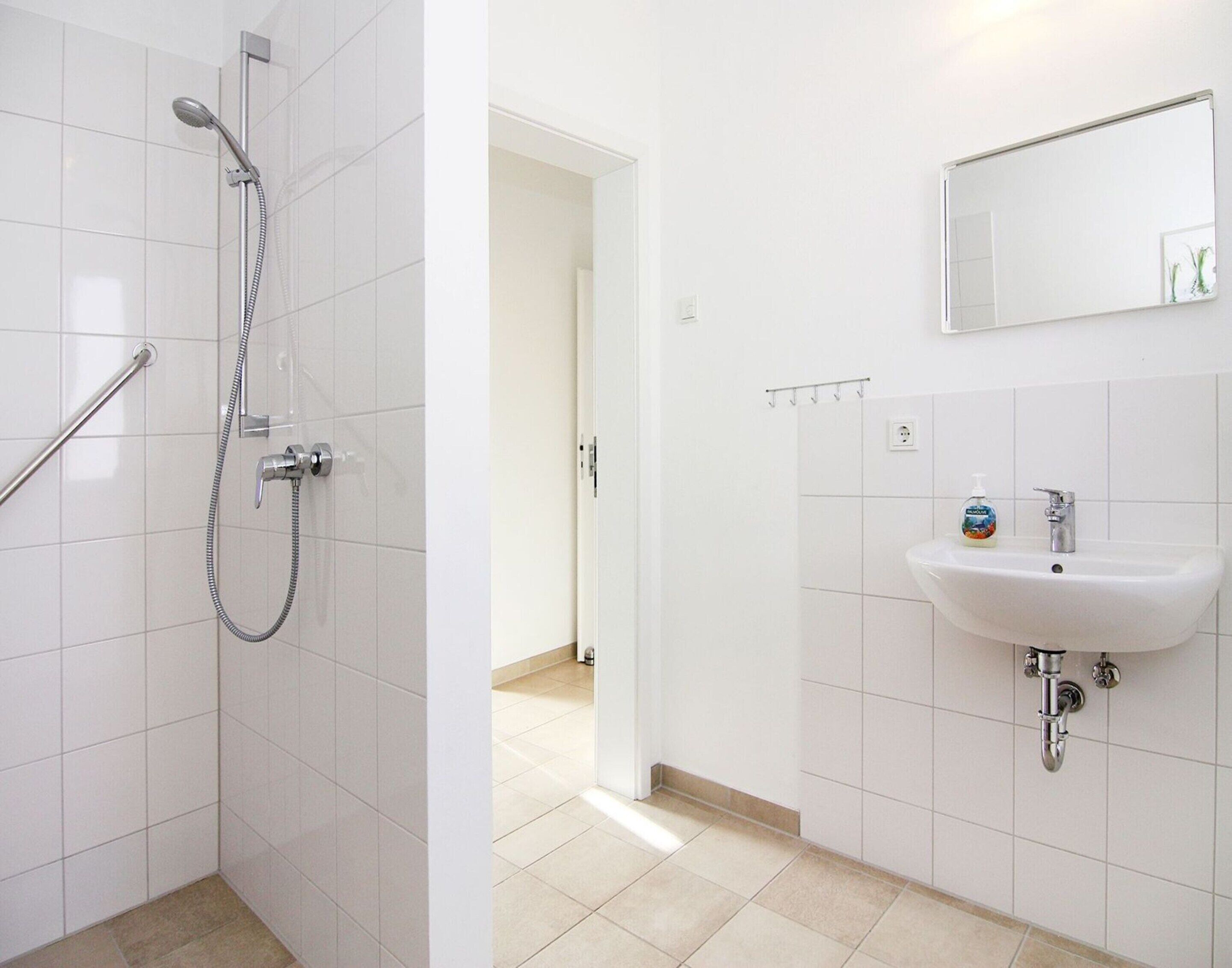 A bathroom with white tiles, a shower with a handheld showerhead, a sink with a mirror above it, and a bottle of soap.