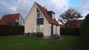 A two-story house with a sloped roof, a small patio with a table and chairs, and a well-maintained lawn.