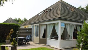A small, single-story building with a tiled roof, a covered patio area with a table and chairs, and a sign indicating a room for rent.