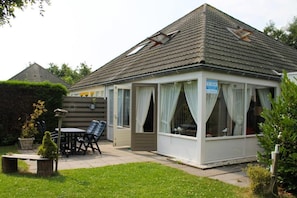 A small, single-story building with a tiled roof, a covered patio area with a table and chairs, and a sign indicating a room for rent.