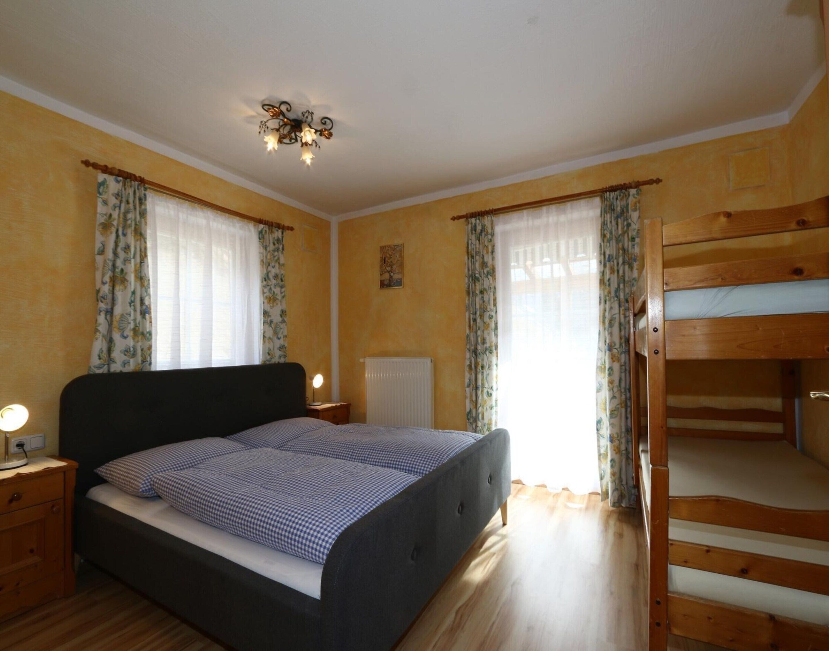 A bedroom with a double bed, a wooden bunk bed, a bedside table with a lamp, and a ceiling light fixture.