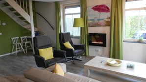 Un salon avec un escalier, deux fauteuils noirs avec des coussins jaunes, un canapé beige, une table basse blanche et un téléviseur posé sur un meuble.