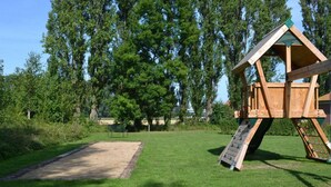 Une structure de jeu en bois, dotée d’un toboggan et d’un filet d’escalade, entourée d’une zone herbeuse et d’arbres.