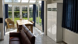 A room with a dining table, chairs, a sofa, a TV, and a view of a garden through large windows.