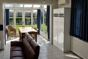 A room with a dining table, chairs, a sofa, a TV, and a view of a garden through large windows.