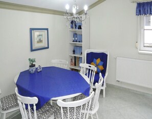 Ein Essbereich mit einem runden Tisch, der mit einer blauen Tischdecke bedeckt ist, umgeben von weißen Stühlen mit gemusterten Kissen. An der Wand befindet sich ein Bücherregal mit verschiedenen Gegenständen, und darüber hängt ein Chandelier.