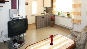 Een woonkamer met een televisie, een bank, een salontafel en een keukenhoek met een raam en gordijnen.