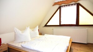 Een slaapkamer met een houten bed, wit beddengoed, een raam met houten kader en een radiator.